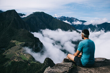 Amazing scenery of high green mountains covered white clouds.Back view of young man dressed in active wear traveler resting and admiring breathtaking nature during hiking wanderlust