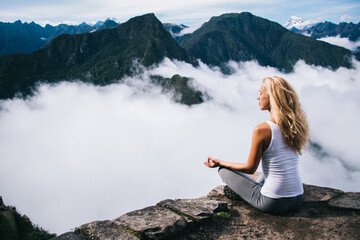 Blonde young woman with blonde hair doing meditation sitting in lotus on edge of rocky hill enjoying relax time of travel.Female tourist doing yoga from high peak mountain enjoying calm of nature