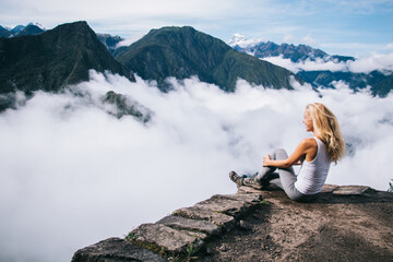 Positive young blonde woman traveler dressed in active wear sitting on rocky hill and admiring breathtaking scenery of high green mountains covered white fog during amazing trip in South America