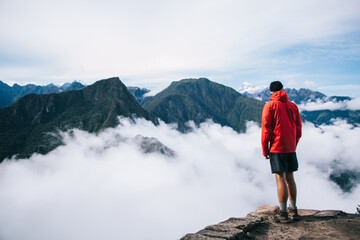 Back view of young man tourist dressed in active wear standing on edge of summit and admiring breathtaking view of green mountains covered white mist.traveler enjoying natural beauty of environment