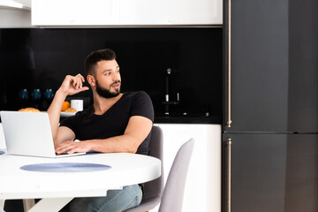 handsome freelancer looking away while sitting near laptop
