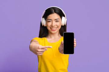 Music Streaming Service. Smiling asian woman with headphones pointing on black smartphone