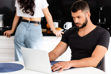 selective focus of handsome freelancer using laptop near girlfriend in kitchen