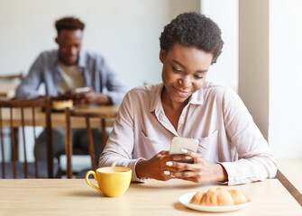 Fototapeta na wymiar Attractive African American girl browsing internet on smartphone during breakfast at cozy city cafe, blank space
