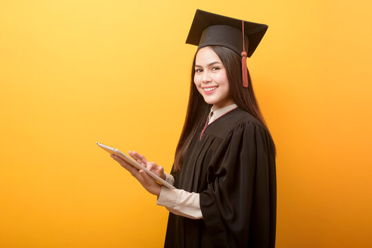 portrait of beautiful woman in graduation gown is holding tablet on yellow background - Powered by Adobe