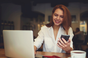 Attractive female international student testing new software for digital devices connecting smartphone and laptop to upgrade system while resting in cafe after lesson using free internet access