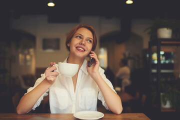 Portrait of attractive smiling female manager having phone conversation with colleague happy to hear about finishing work on successful startup project while drinking coffee on break in cafeteria