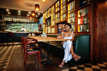 African american woman, retro hairstyle in white dress at restaurant with glass of wine.