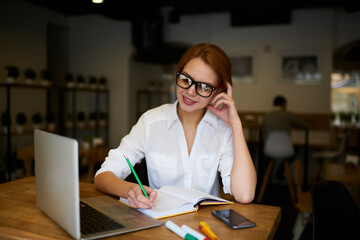 Fototapeta premium Charming talented female student in spectacles gathering information from websites on laptop connected to wireless internet and making coursework project while recording notes to textbook indoors
