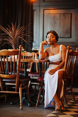 African american woman, retro hairstyle in white dress at restaurant with glass of wine.