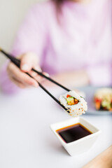 Close up of woman holding sushi roll with chopsticks above bowl full of soy sauce. Healthy traditional japanese meal concept.