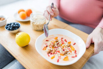 Close up of young pregnant woman sitting on bed and eating granola with cereals and yogurt.