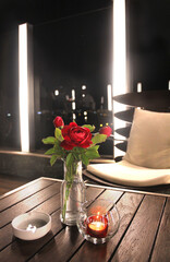 Coffee table in a night cafe. Red rose in a glass bottle, a candle in a transparent glass and a white ashtray on a wooden surface. Modern and stylish furniture on a black background with backlight.