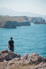Fototapeta na wymiar young man standing on the cliff and watching beautiful coast line near Llanes, Turquoise calm water of Atlantic ocean, sunny weather