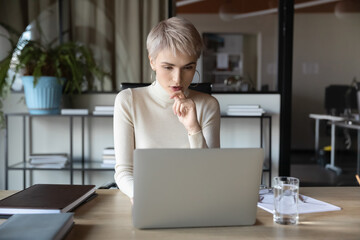 Concentrated businesswoman sitting at desk in contemporary office room looking at laptop screen...