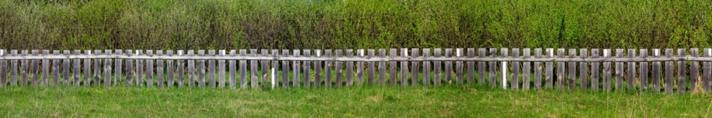 Panorama of a wooden fence on a green background. Grey wooden fence in summer, against a background of green bushes. Fence around a garden or private house