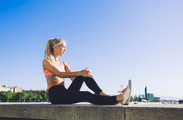 Tired female jogger taking break after fitness training while sitting on pier against blue sky background with copy space area for advertising, young sports woman enjoys resting after workout outdoors
