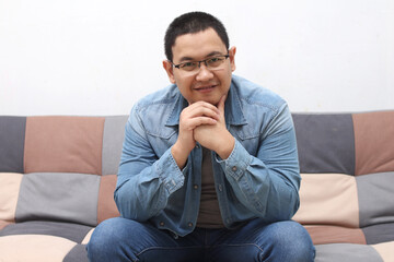 Handsome young Asian man sitting at home on sofa, smiling happy, looking at camera