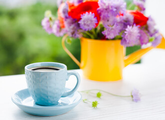 Obraz na płótnie Canvas Cup of coffee and beautiful bouquet of bright wildflowers in yellow watering can on white wooden background.