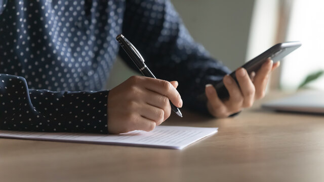 Close up student sit at desk hold pen writing essay look at cell screen search helpful info prepare for university exams. Businesswoman financier working use app, reading latest financial news concept