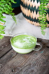 Glass cup of matcha latte on wooden table with room plant on background.