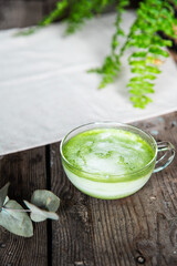 Glass cup of matcha latte on wooden table with room plant on background.