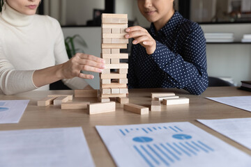 Multiracial businesswomen play build wood block stacking game, financial stats report on desk close up. Cooperation, company stability, reach aim effort, business strategic thinking management concept