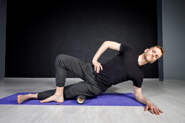 Young man is stretching his outer 
hip muscles with the help of yoga roller.