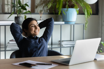 Carefree indian employee resting after busy fruitful workday leaned on office chair puts hands...