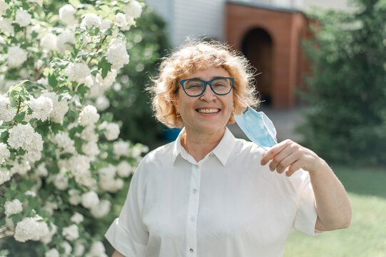 Happy Old Woman Takes Off The Mask And Sniffs A Blossoming Apple Tree. A Female Pensioner Enjoys Fresh Air On A Warm Summer Day In The Garden. Quarantine Is Over. The End Of The Coronavirus Pandemic.