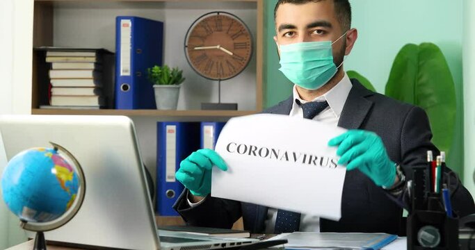Young businessman sitting at the table, holding Coronavirus word and looking to the camera with medical protective mask and gloves