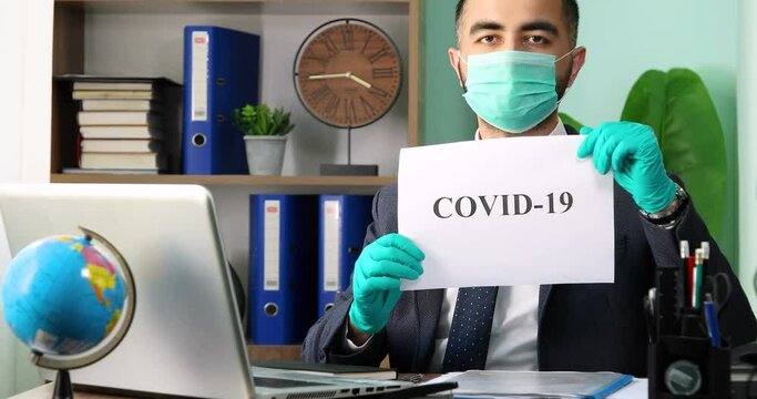 Young businessman sitting at the table, holding COVID-19 word and looking to the camera with medical protective mask and gloves