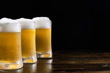 Three ice-cold beer mugs on wooden base on black background with copy space. Sselective focus.