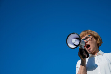 Angry senior woman in glasses shouting into a megaphone on the outside. Annoyed pensioner yells...