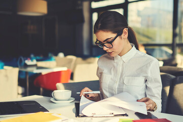 Skilled professional female administrative manager of coffee shop checking documentation for month...
