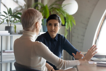 Indian and caucasian businesswomen negotiating sit at desk in office. Lawyer consulting client during formal meeting. Job interview and hiring process. Diverse colleagues discussing project concept - obrazy, fototapety, plakaty