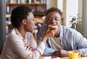 African American girl feeding delicious sweet tartlet to her boyfriend at coffee shop