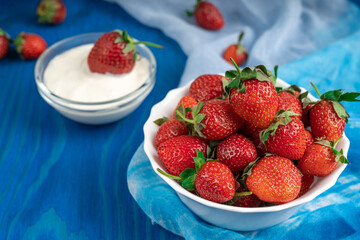 White bowl full of ripe strawberries on blue wooden surface against bowl of yogurt