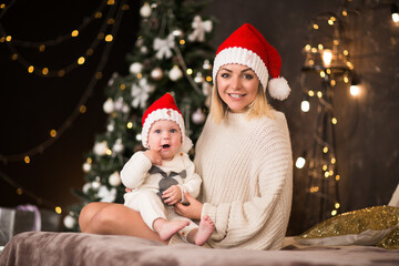 Fototapeta na wymiar Young woman in red Santa's cap posing with little baby boy in red cap against Christmas tree's lights.