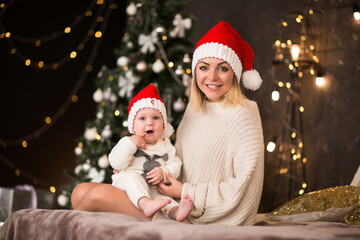 Young woman in red Santa's cap posing with little baby boy in red cap against Christmas tree's lights.