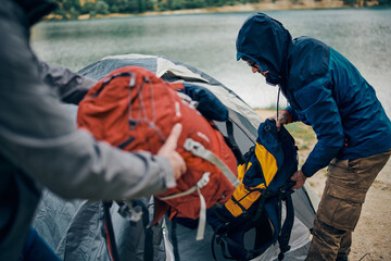 Fototapeta premium Two men on camping trip putting backpacks into tent by the lake on rainy and cold weather.