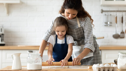 Obraz na płótnie Canvas Smiling mother teaching little daughter to roll out dough standing at table in kitchen. Happy caring mum and adorable girl using rolling pin on wooden board, preparing dinner, cooking flour.