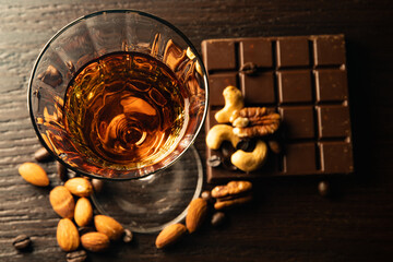 Cognac wine glass with amber cognac, nuts lying near the wine glass, coffe grains, chocolate bar on...