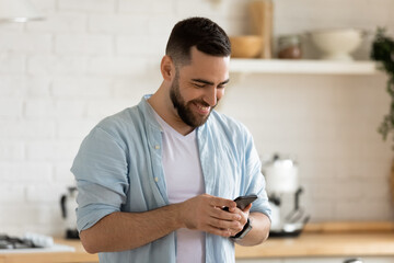 Close up happy handsome man reading good news on smartphone in kitchen at home. Smiling bearded guy...