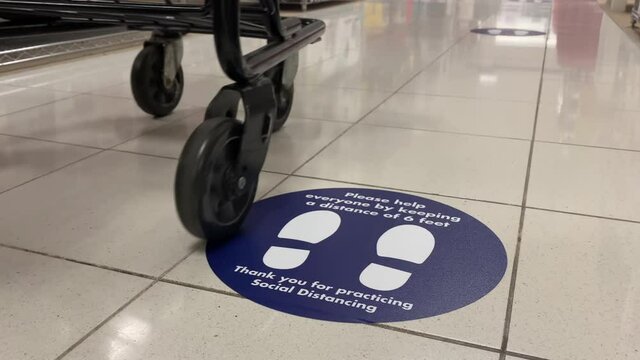 A low angle view of a social distance reminder sticker on the floor of a department store as an elderly woman pushing a shopping cart walks past.	