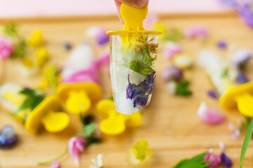 Hand holding floral Ice Pop with summer wildflowers on background of ice cubes and fresh summer flowers on wood. Hello summer concept