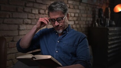 Mature man in his 50s reading book at home sitting in dark living room. 