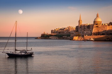 View on Malta walls during sunset with visible moon and a yacht in the foreground.  - obrazy, fototapety, plakaty