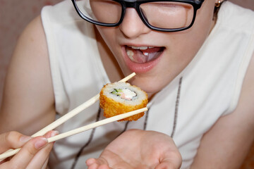 A girl with glasses and piercings in her tongue puts a sushi roll in her mouth with chopsticks.