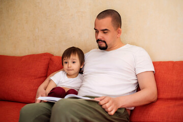 Father reading book to toddler son, child is looking away and thinking. Dad and little son sitting on sofa and reading fairy tales. Father and son spending good time together at home